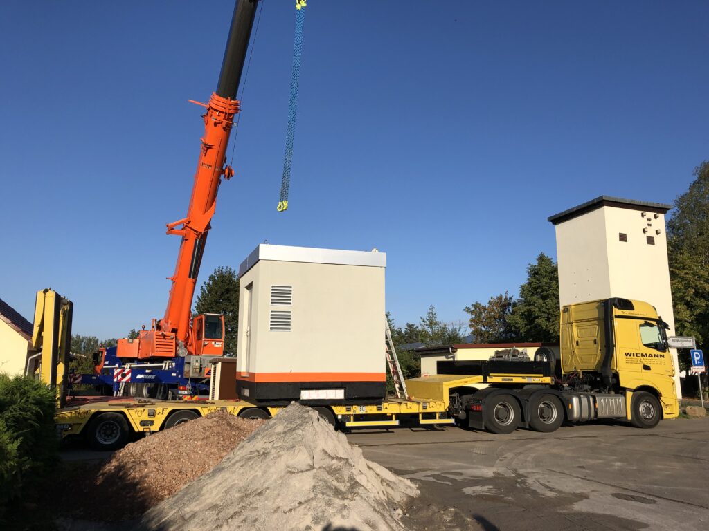 Ein Schwerlastkran hebt eine POP-Station von einem gelben LKW-Anhänger, im Hintergrund sind Wohnhäuser und Bäume sichtbar.