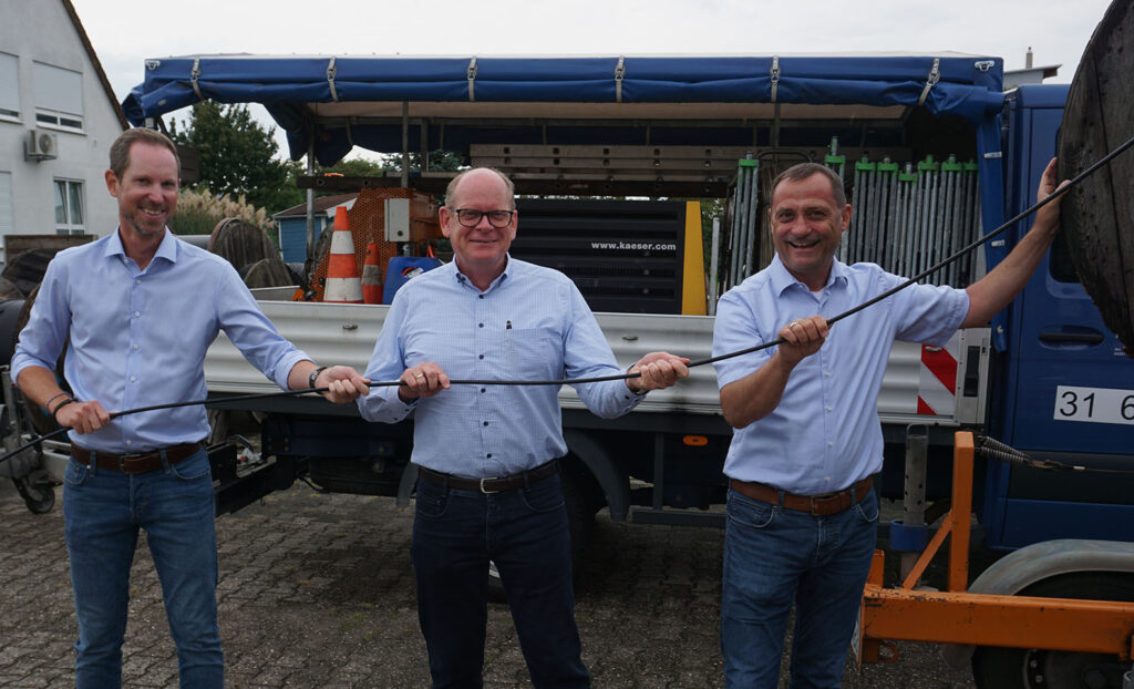 Drei Männer bei symbolischer Handlung zum Glasfaserausbau, halten gemeinsam ein Kabel vor einem Montagefahrzeug.
