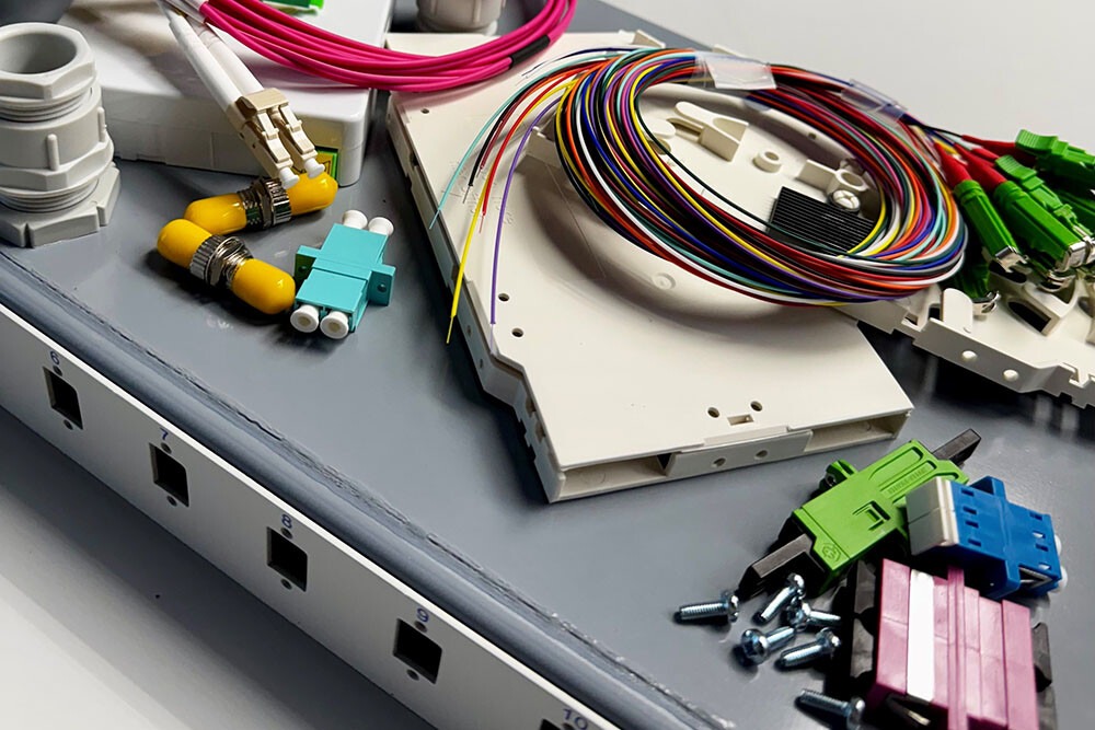 Unteschiedliches Breitband-Zubehör wie Spleisskassetten, LWL-Stecker und Kupplungen, LWL-Patchkabel auf grauer Arbeitsfläche verteilt.